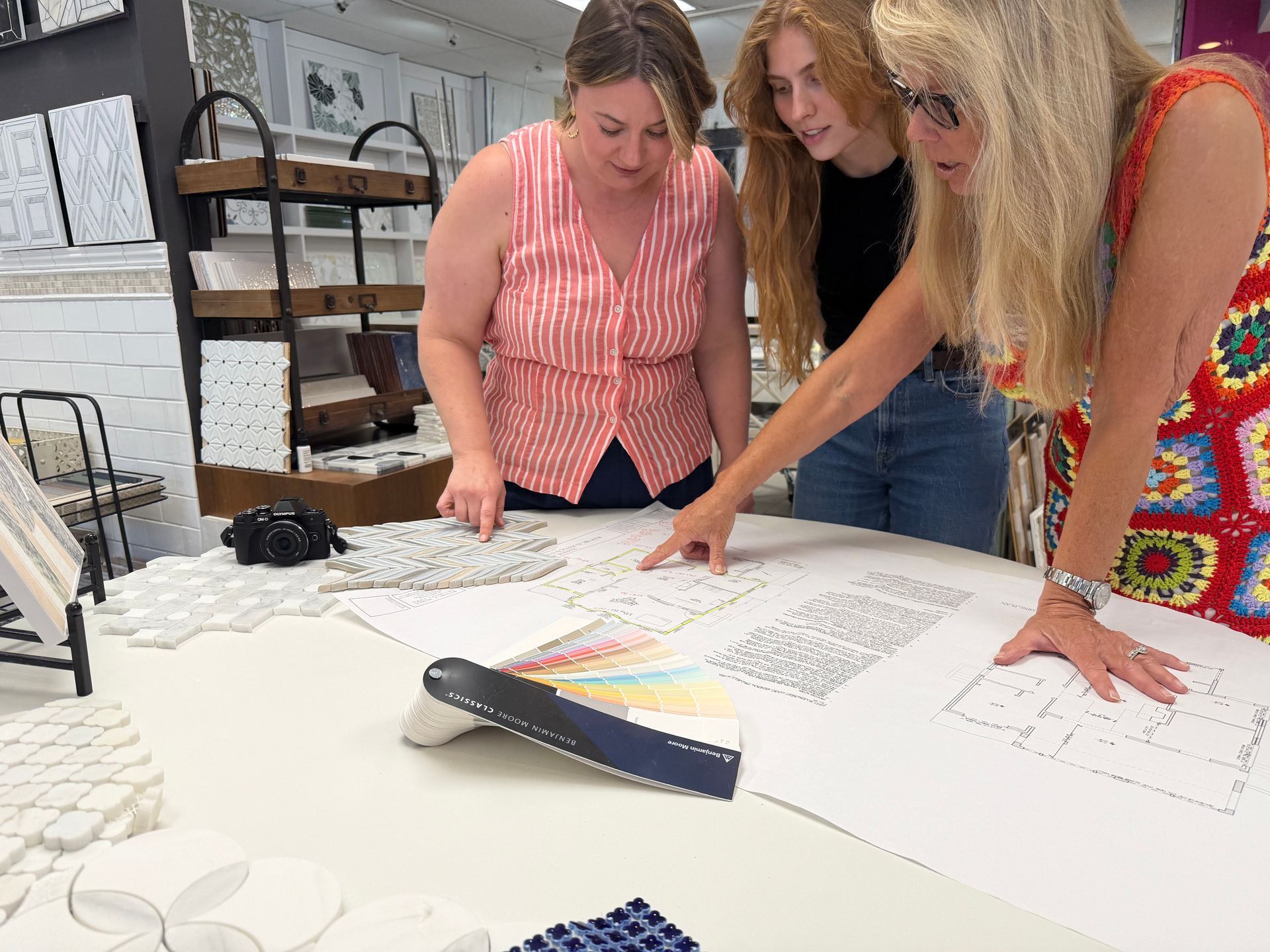 Three people lean over a large table reviewing papers and a colorful folded sample in a bright workspace.