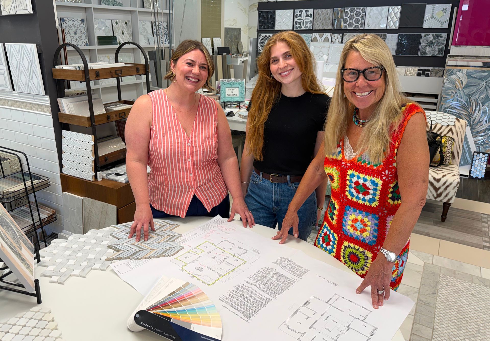 Three women smiling behind a table of design sketches and samples in a bright studio.