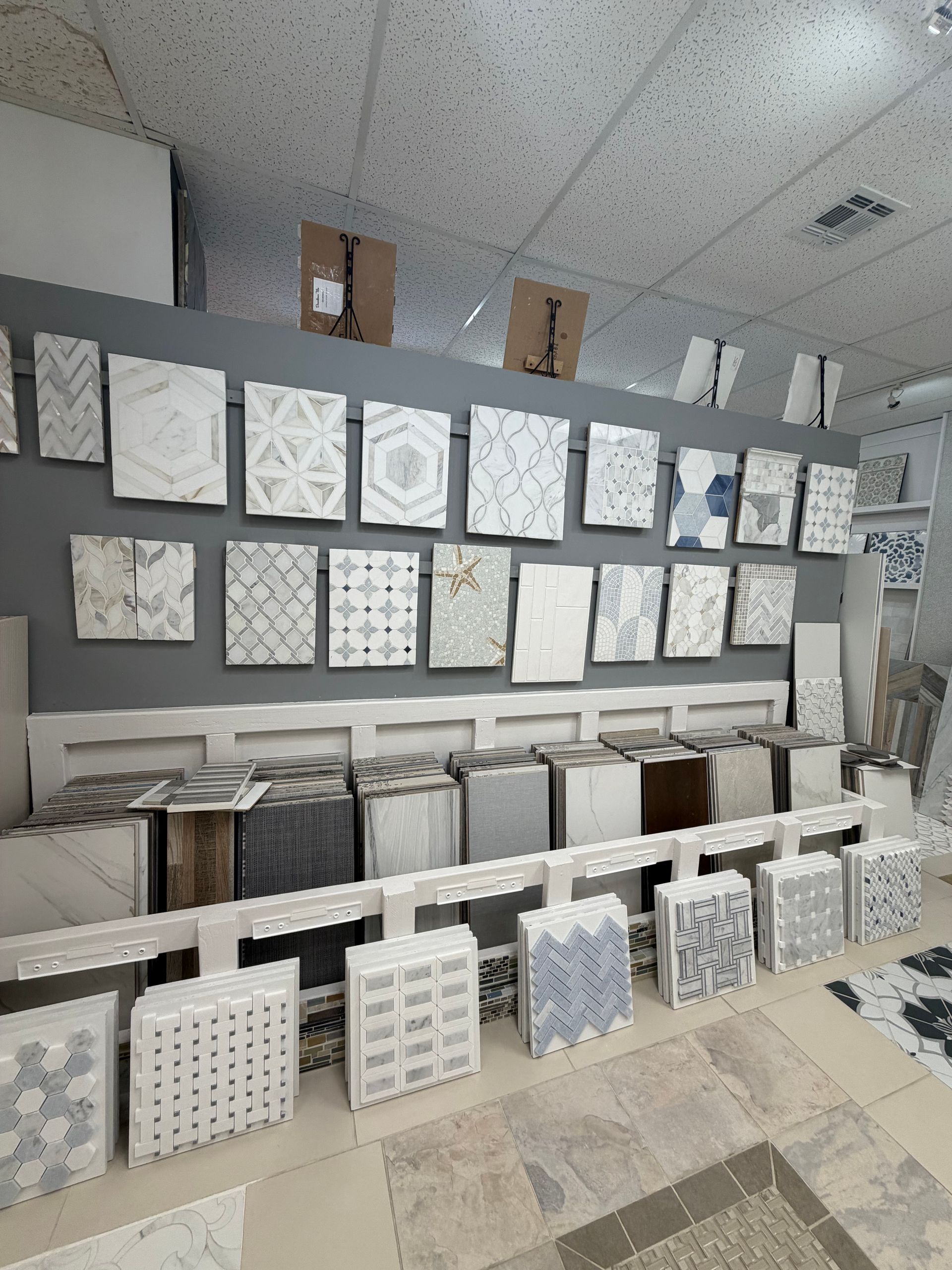 Display of white tile samples and patterned panels in a showroom, arranged on wall shelves and floor stands.