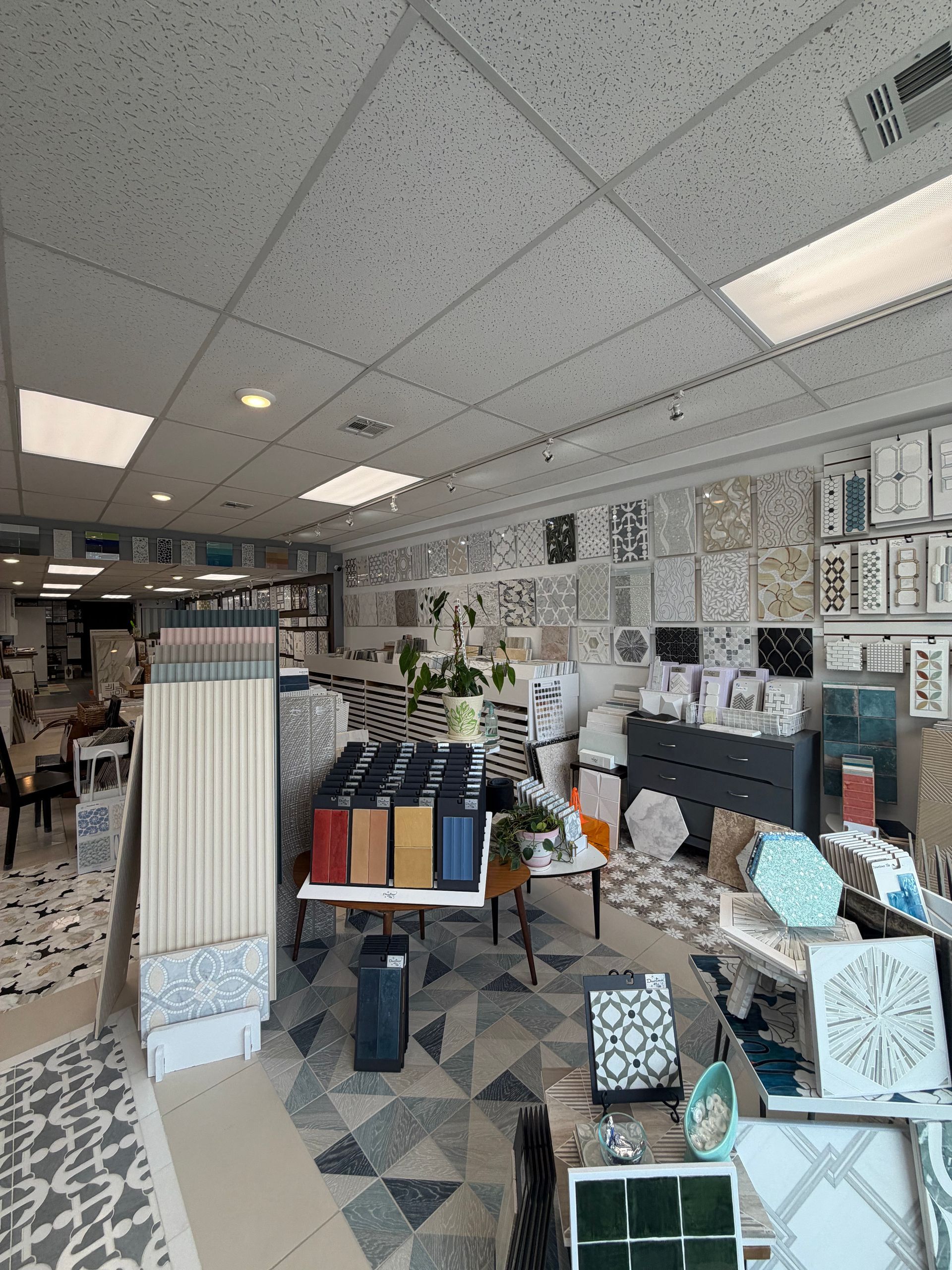 Bright showroom with tiled ceiling, patterned rugs, and display tables of home decor items.