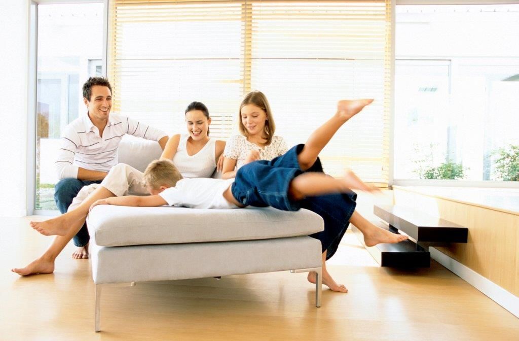 Family laughing on a couch as two children play and kick their legs, in a modern living room.