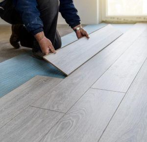Person installing light-colored wood floor planks over blue underlayment.