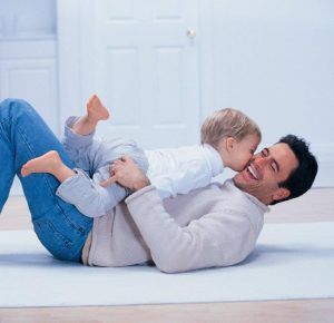 Father lying on floor, playing with a child, child kissing his cheek, both smiling.