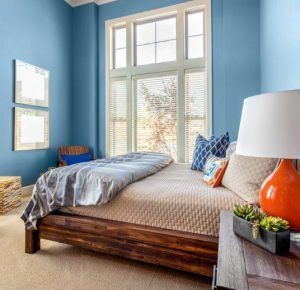 Bedroom with blue walls, wooden bed, large window with blinds, orange lamp, and two square mirrors.