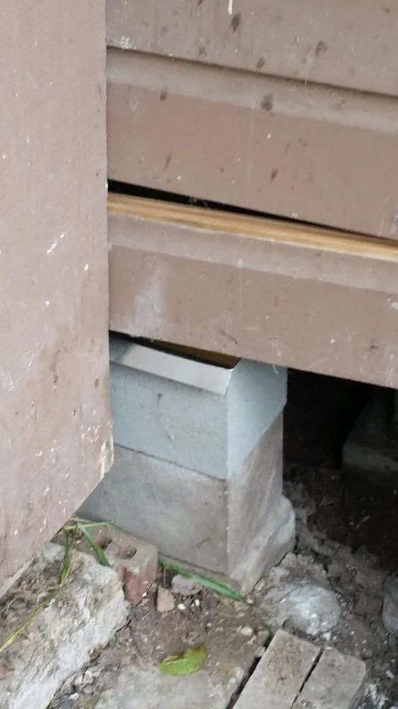 Damaged house foundation with a concrete block supporting a brown siding panel above a crawlspace opening