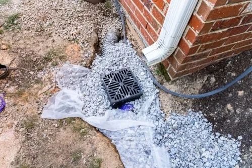 Broken drain pipe beside a brick house, with gravel and plastic sheeting around a storm drain.