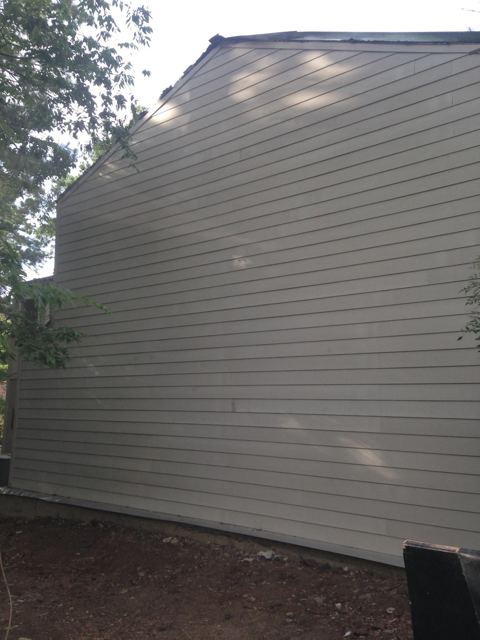 Side view of a beige vinyl-sided house wall with trees and a shaded yard.