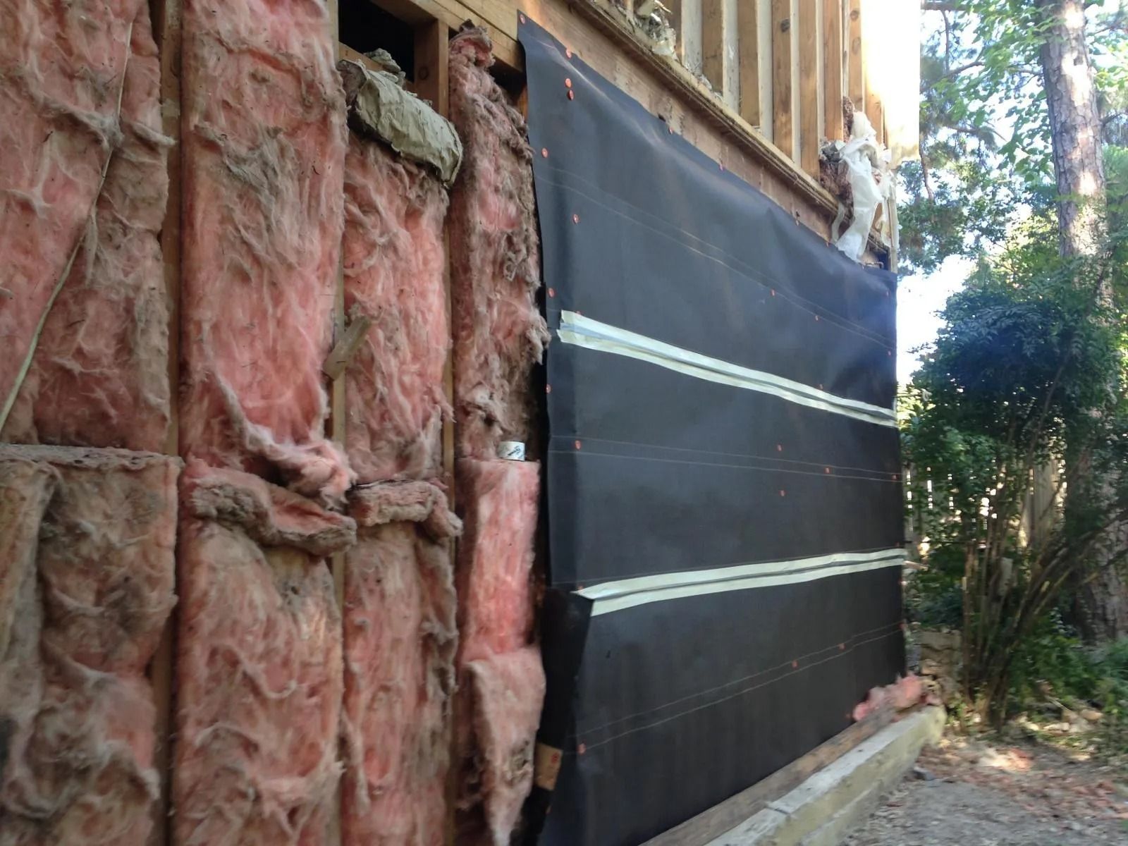 Outdoor construction site with black tarp-covered wall beside a red stone wall and trees.