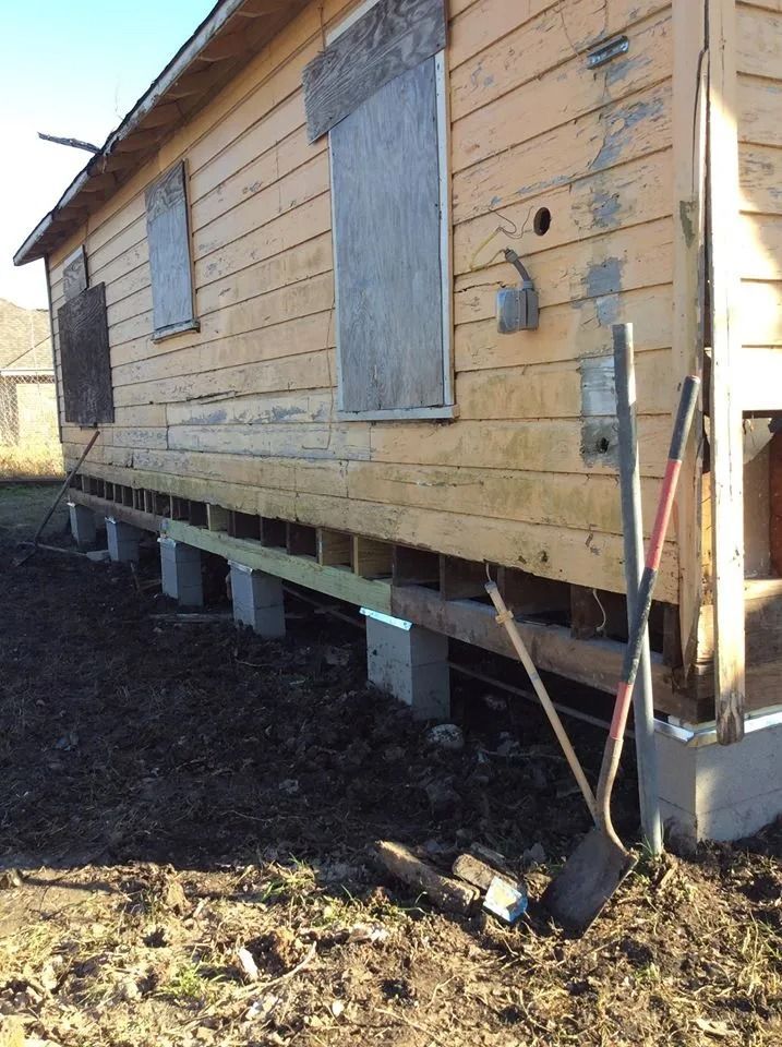 Old weathered wooden house on blocks with boarded windows, tilted in a muddy yard
