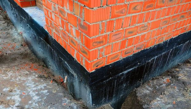 Corner of a red brick building with a black waterproofing strip at the base on a concrete surface