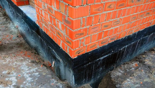 Corner of a red brick building with a black waterproofing strip at the base on a concrete surface