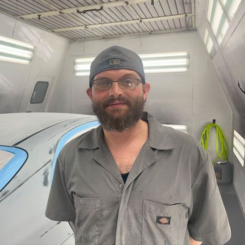 A man with a beard and glasses is standing in front of a car in a paint booth.
