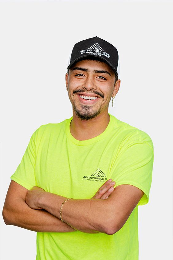 A man wearing a yellow shirt and a black hat is standing with his arms crossed and smiling.