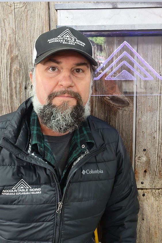A man with a beard and a hat is standing in front of a wooden wall.