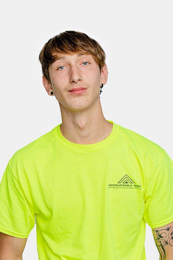 A young man wearing a neon yellow t-shirt is standing in front of a white background.