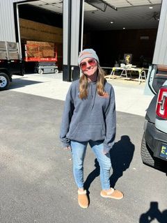 Person in sunglasses and gray sweater standing outside a garage in sunlight.