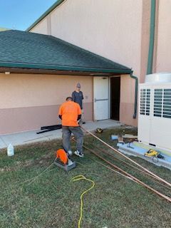 Workers installing pipes near a building and HVAC unit on a grassy lawn.