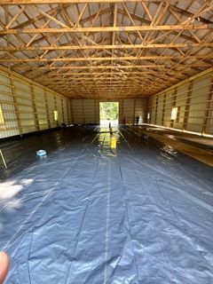 Empty barn interior with exposed wooden trusses and a dark tarp-covered floor, open doorway at the far end.