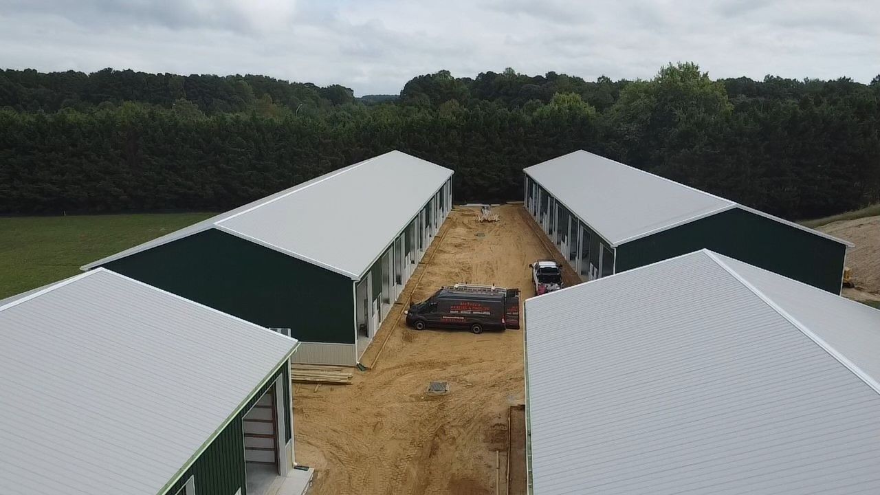 Construction site with several white-roofed buildings around a dirt lane, bordered by trees.