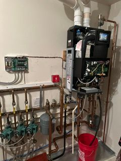 Utility room with wall-mounted boiler, exposed pipes, valves, and a red bucket on the floor