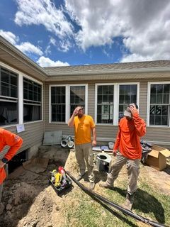 Three workers standing in a backyard patio area, one in orange shirt and two in orange tops, with tools nearby.