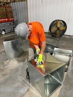 Worker in orange shirt and hard hat bending over a metal duct in a workshop.
