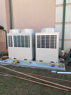 Two large white rooftop HVAC units beside a building wall with tools and pipes on the ground