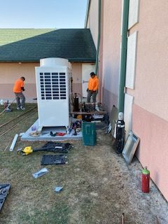 Workers install equipment beside a building wall, with tools and a large HVAC unit on the ground.