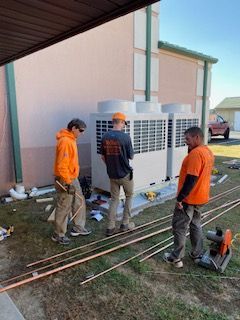 Three workers in orange and dark shirts stand beside an outdoor HVAC unit and tools under a covered area.