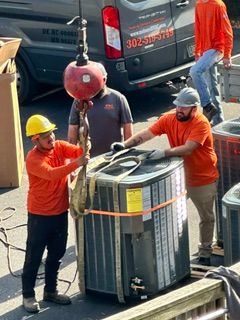 Workers in hard hats move a large HVAC unit near a service truck.