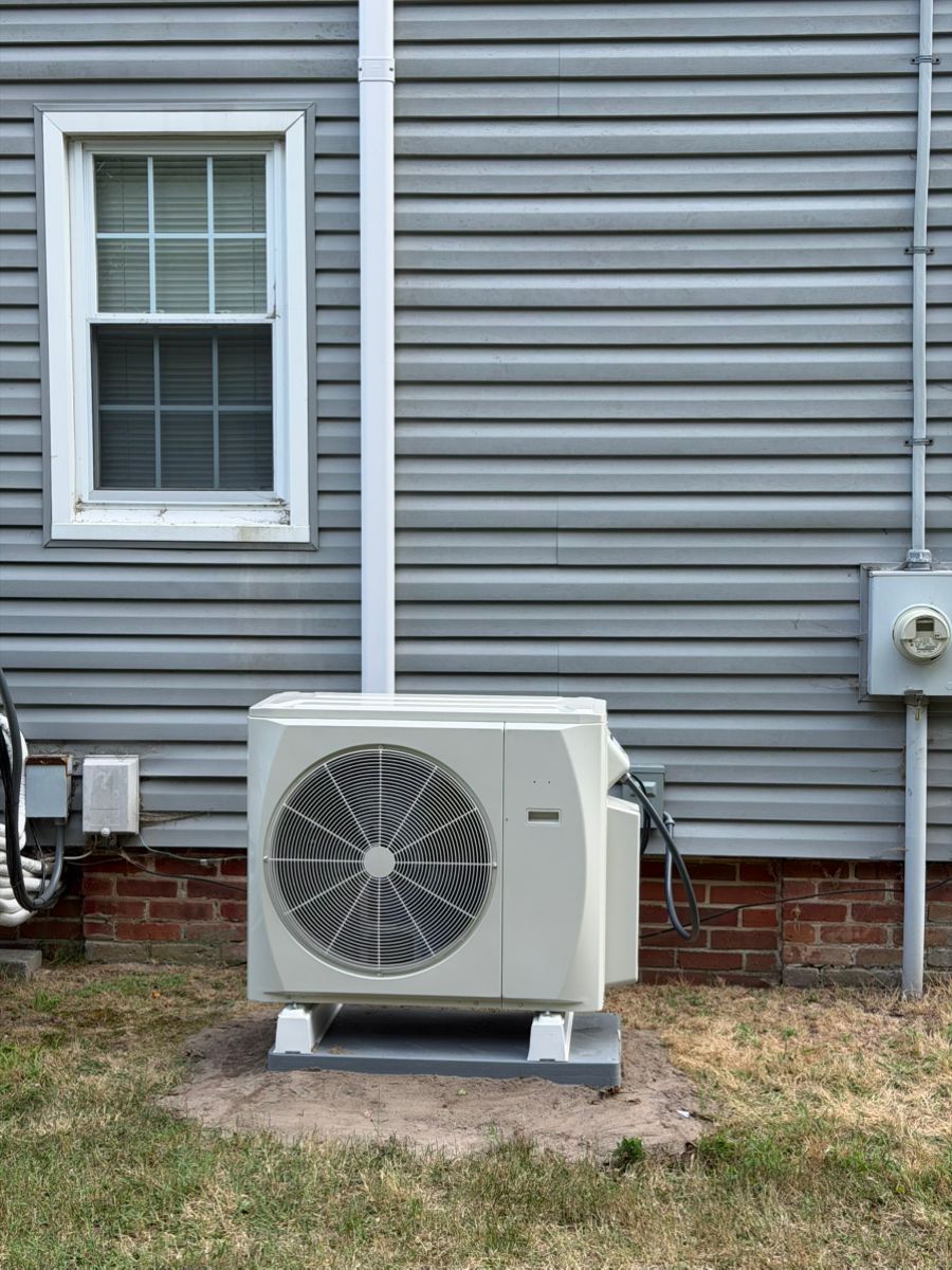 Outdoor air conditioner unit beside a gray house with vinyl siding and a small window.