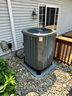 Outdoor air conditioner unit beside a house window, surrounded by rocks and a wooden deck railing.