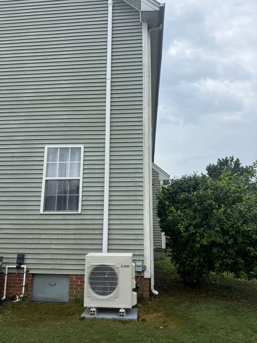 Side of a gray house with a window, downspout, and outdoor HVAC unit beside a grassy yard.
