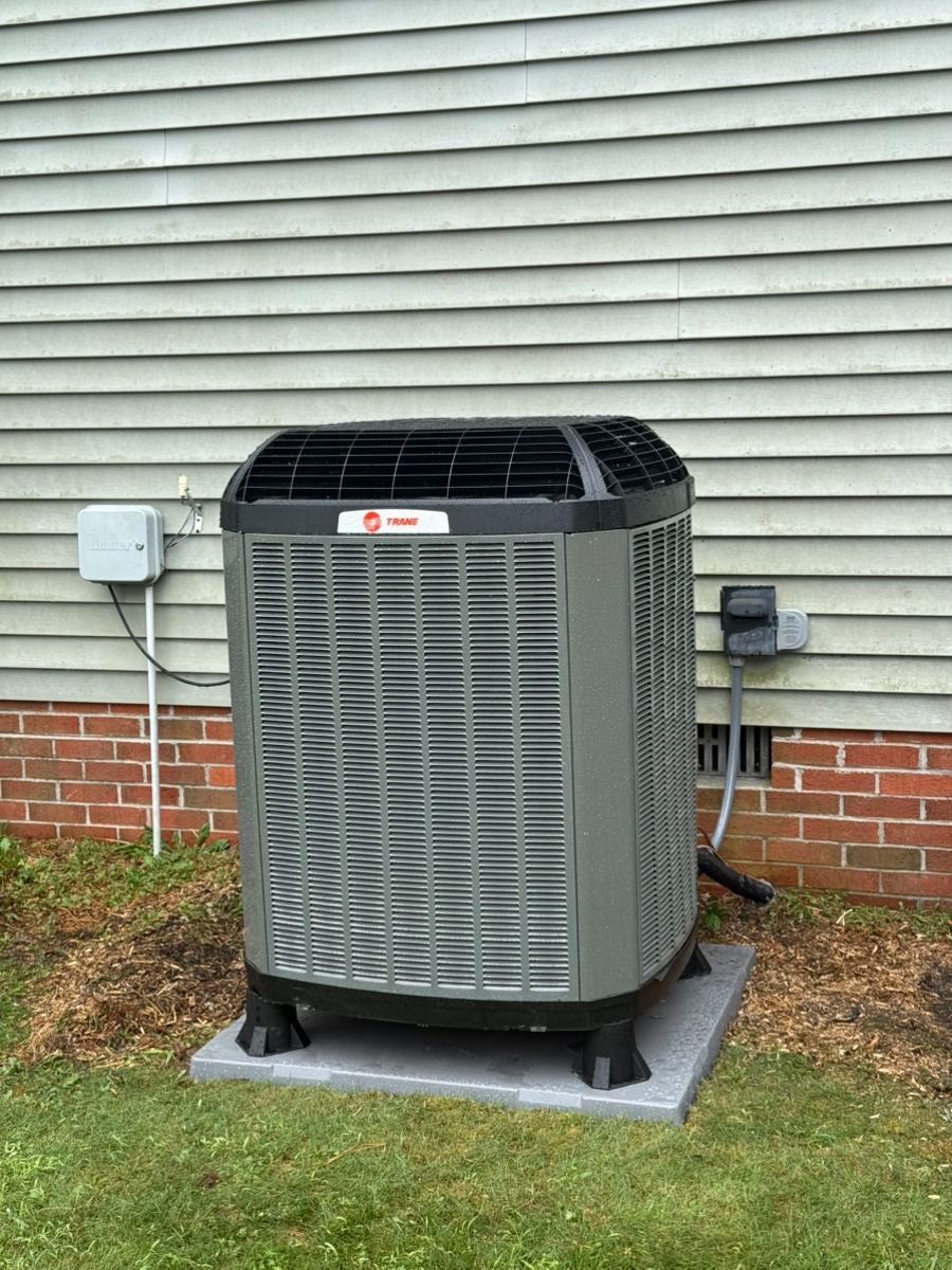 Outdoor central air conditioning unit beside a house wall and brick foundation, on a concrete pad.