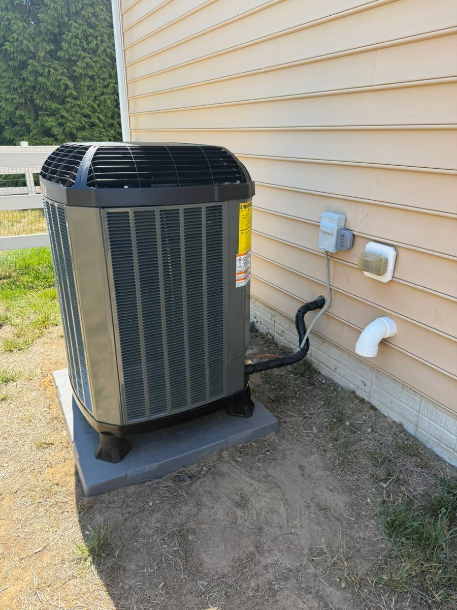 Outdoor air conditioner unit beside a house wall with utility pipes and tan siding