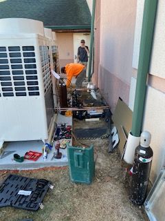 Worker in orange pants repairing an outdoor HVAC unit beside a house
