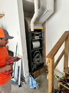 Worker inspecting furnace ductwork in a utility closet with exposed pipes and a staircase nearby