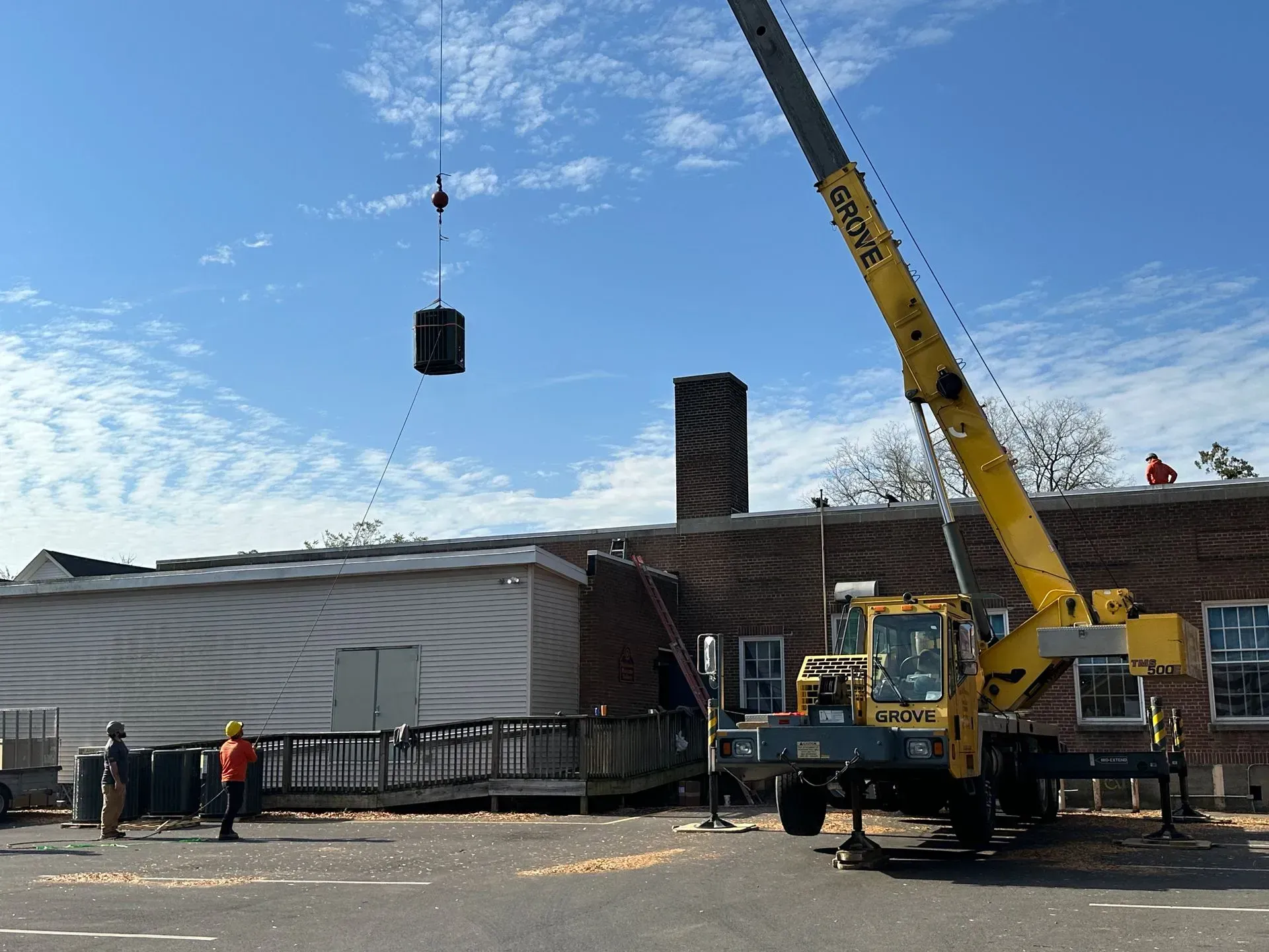 Crane lifting a dark cylindrical load beside a large building under construction, with workers nearby.