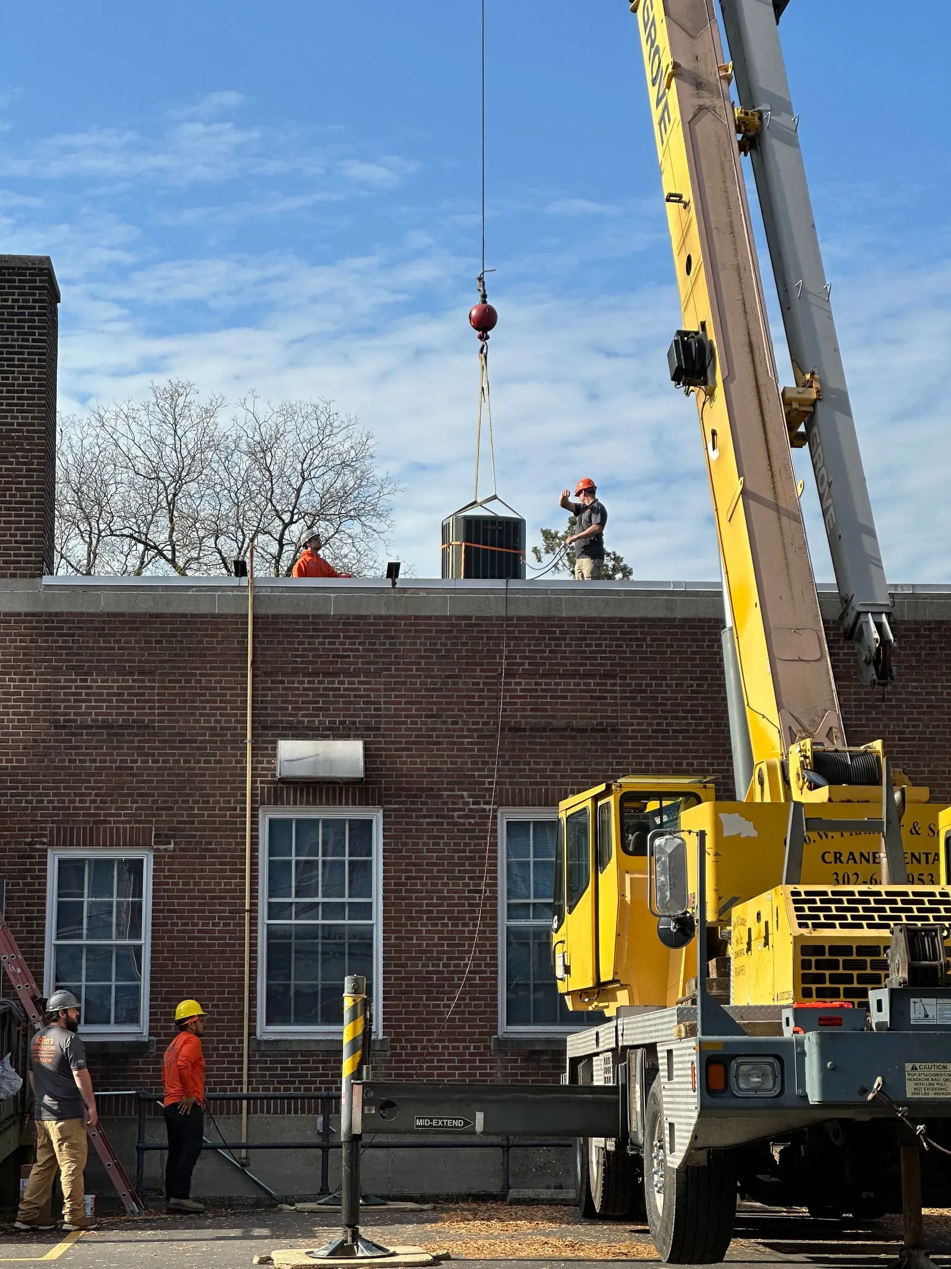 Yellow crane lifting workers onto a brick rooftop beside a building under a blue sky