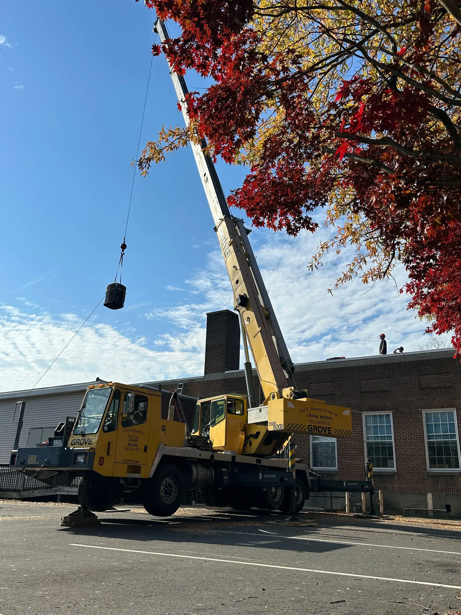 Yellow crane lifting a black bucket beside a building under a blue sky, with red autumn leaves framing the scene