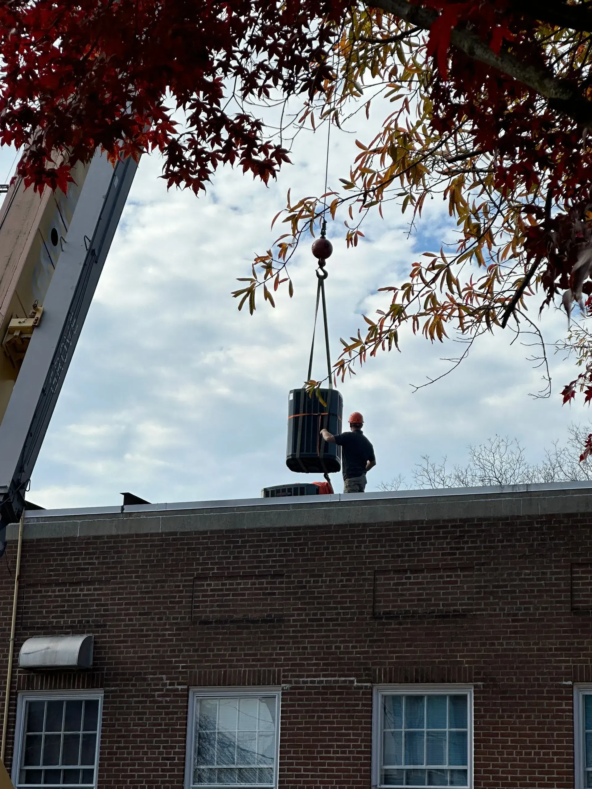 Worker on a roof lifts a black object with a crane beside a brick building.