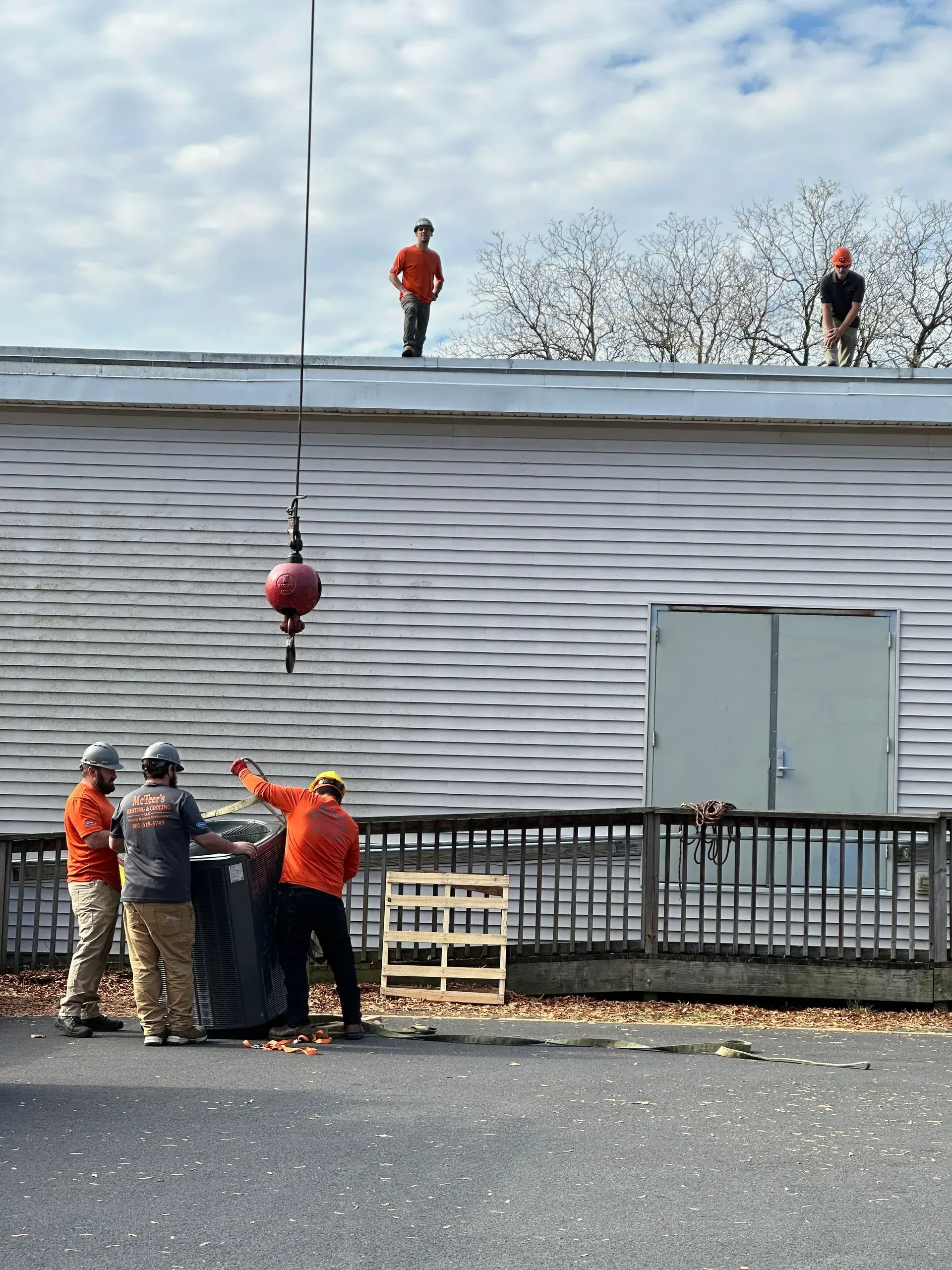 Workers on a roof and ground crew below lifting materials beside a gray building