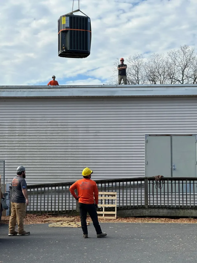 Workers guide a black dumpster being lifted over a gray building roof by a crane.