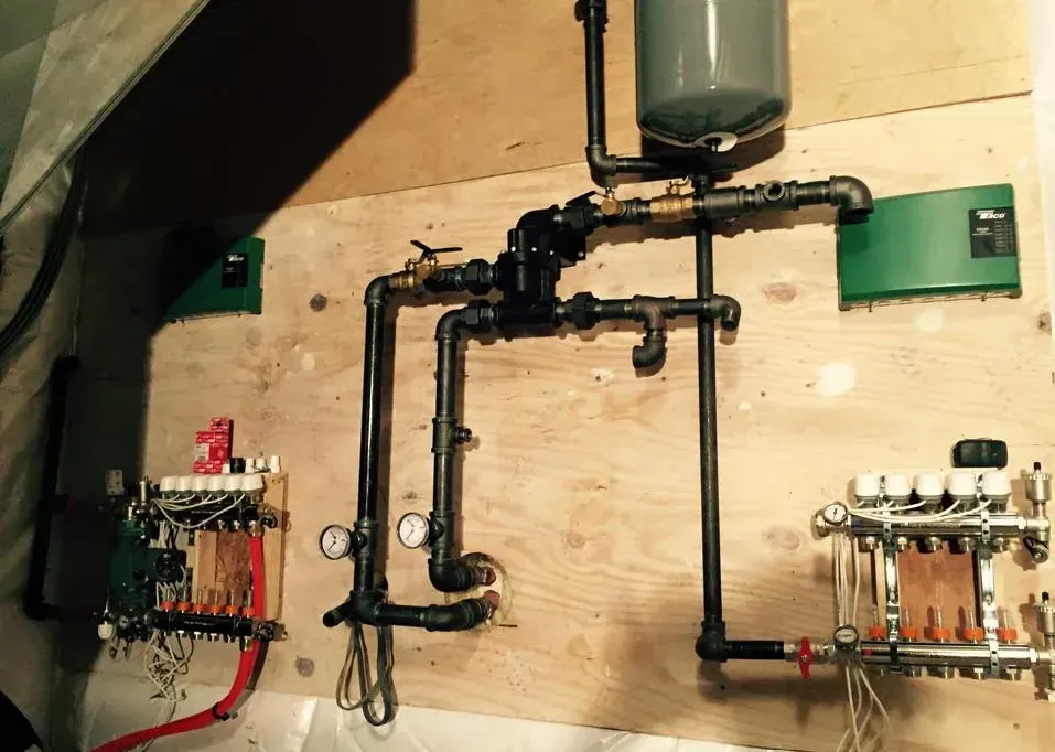 Wall-mounted plumbing manifold with black pipes and red/white valve assemblies in a utility room