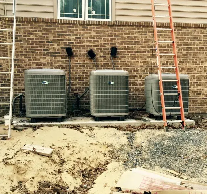 Three outdoor HVAC units beside a brick house, with a ladder leaning nearby and dirt ground in front.