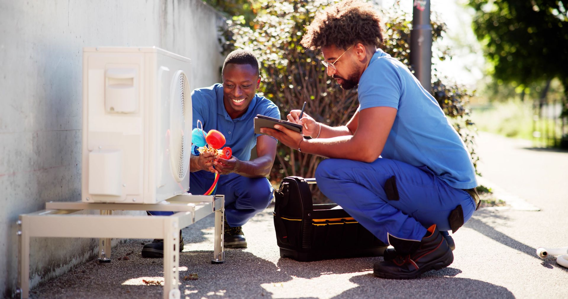 Two people crouch by a small outdoor machine, inspecting colorful parts in bright sunlight.