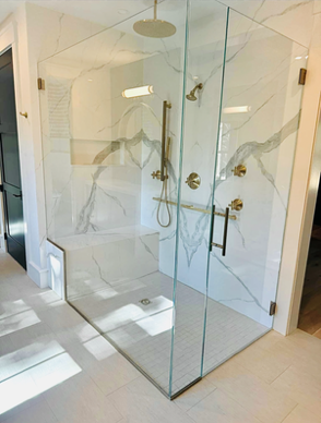 Spacious, corner shower with glass walls and white marble. Features a bench, multiple showerheads, and gold fixtures.