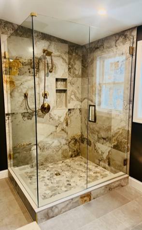 Glass-enclosed shower with marble-patterned tile walls and floor. Gold fixtures, small window, built-in shelf.