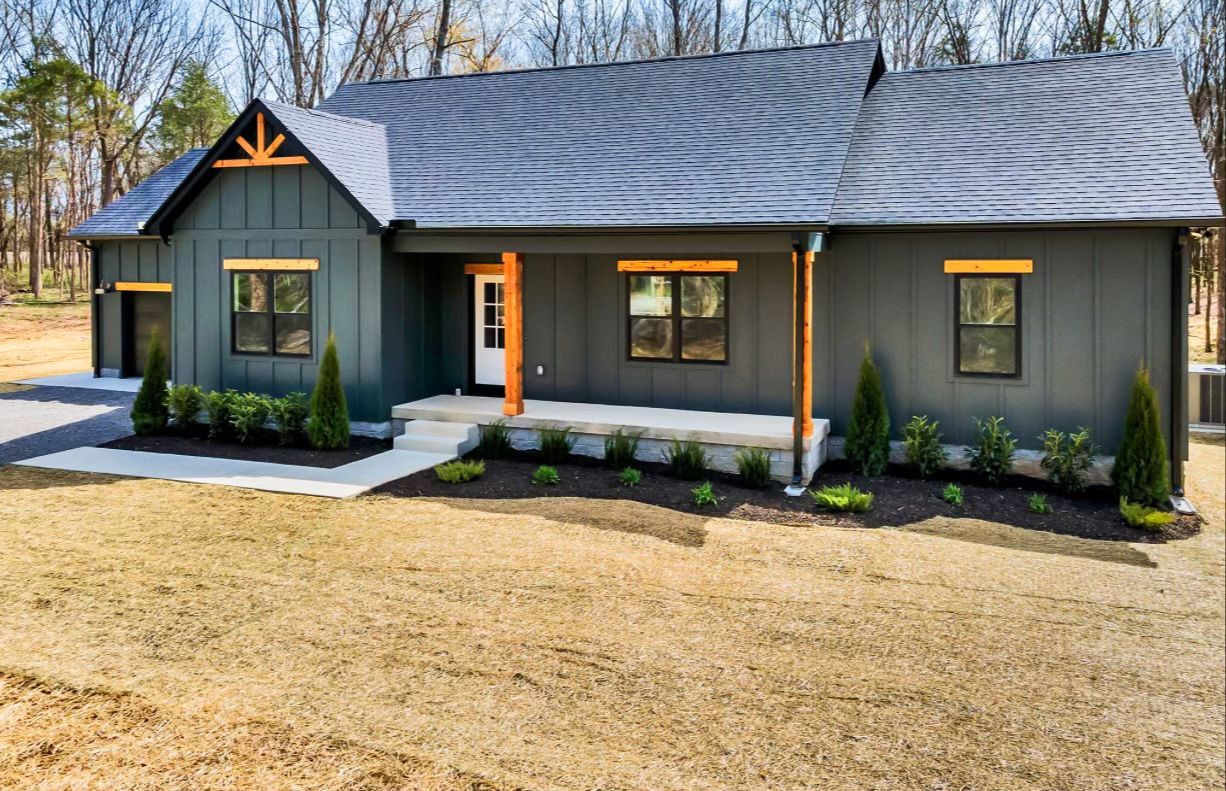 A large gray house with a porch and a garage is sitting on top of a dirt field.