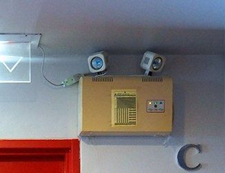 Wall-mounted tan control box with two small security cameras above a red doorway in a hallway.